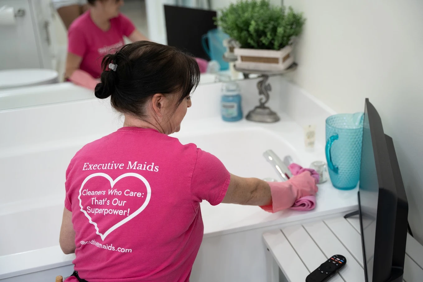 An Executive Maids Cleaning Professional wiping down a bathtub in a Boynton Beach home, wearing a shirt reading Cleaners Who Care: That's Our Superpower
